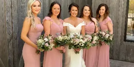 Bride and bridesmaids holding custom floral bouquets in front of a wooden barn wall.