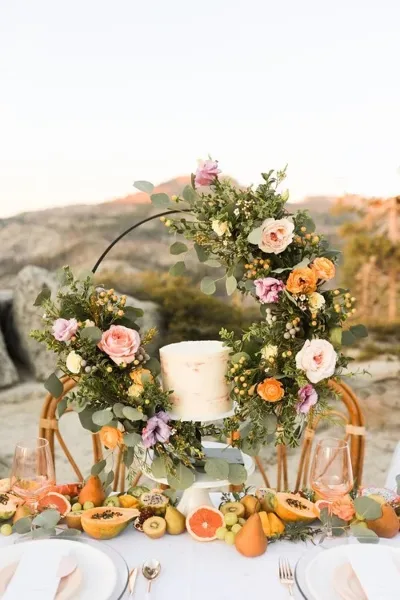 A wedding reception table centerpiece featuring a floral arch with roses and greenery surrounding a white cake.