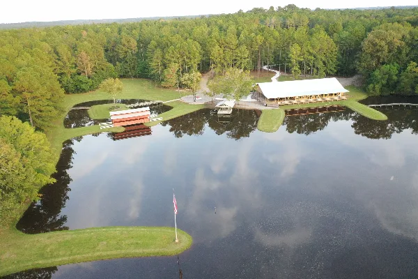 Aerial view of a rural event venue featuring a large white-roofed building and a red barn situated on the shore of a large lake surrounded by pine forest.