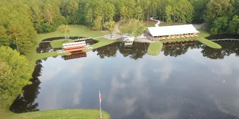 Aerial view of a rural event venue featuring a large white-roofed building and a red barn situated on the shore of a large lake surrounded by pine forest.