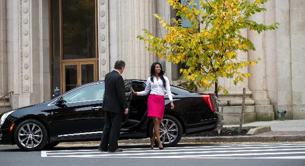 A professional driver in a suit opens the door of a black luxury sedan for a female passenger on a city street.