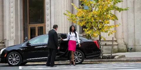 A professional driver in a suit opens the door of a black luxury sedan for a female passenger on a city street.