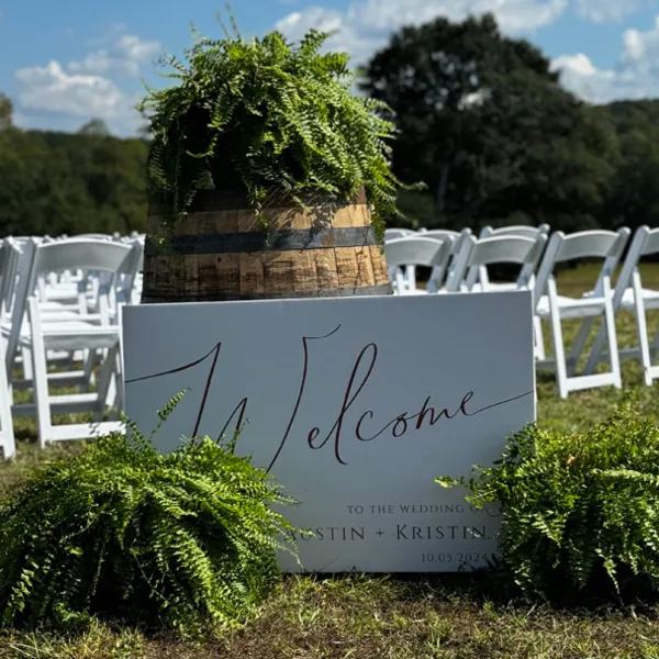An outdoor wedding ceremony setup featuring white chairs and a welcome sign with greenery.