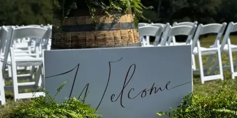 An outdoor wedding ceremony setup featuring white chairs and a welcome sign with greenery.