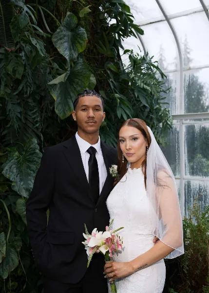 A wedding couple posing in a greenhouse with lush greenery, the groom in a black suit and the bride in a white lace dress and veil holding a bouquet.