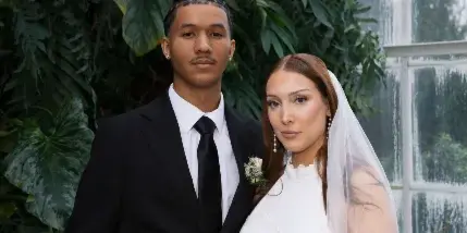 A wedding couple posing in a greenhouse with lush greenery, the groom in a black suit and the bride in a white lace dress and veil holding a bouquet.