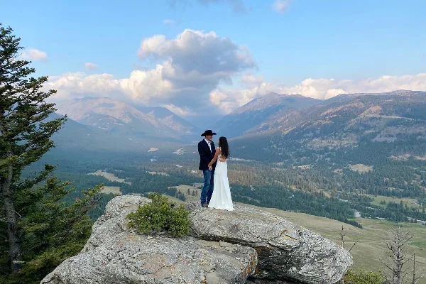 A bride and groom standing on a rocky ledge overlooking a mountain valley.
