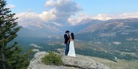 A bride and groom standing on a rocky ledge overlooking a mountain valley.