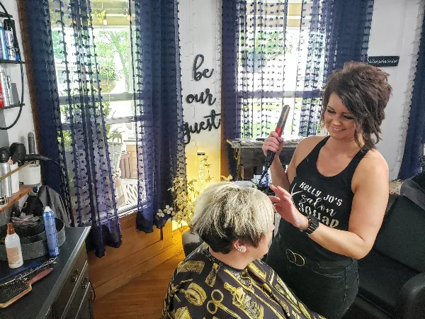 A hair stylist wearing a 'Kelly Jo's Salon' shirt styling a client's hair in a salon setting.