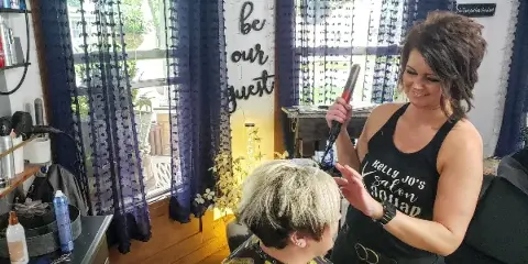 A hair stylist wearing a 'Kelly Jo's Salon' shirt styling a client's hair in a salon setting.