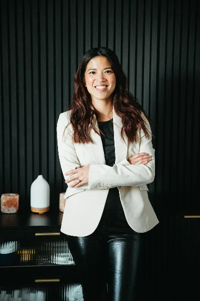 A smiling woman in a white blazer standing in a studio with makeup brushes and spa products on a shelf behind her.