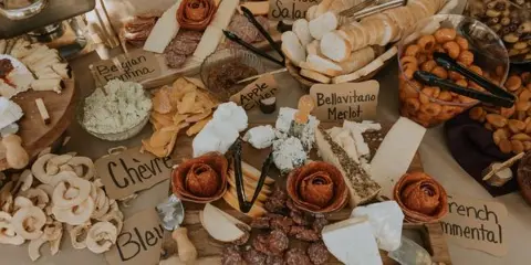 A close-up view of a catering spread featuring a cheese and charcuterie board with various labeled cheeses, meats, and accompaniments.