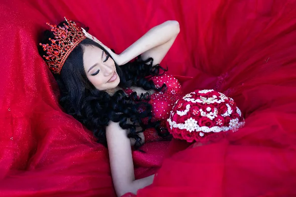 A young woman in a red gown and tiara holding a red bouquet, photographed in a professional studio setting.
