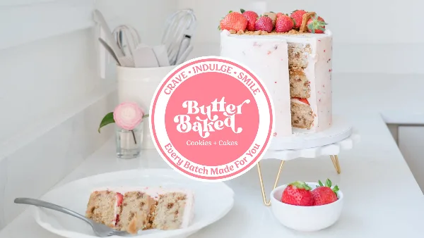 A strawberry-topped custom cake on a white stand with a plated slice and a small bowl of strawberries on a kitchen counter.