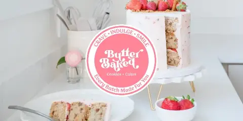 A strawberry-topped custom cake on a white stand with a plated slice and a small bowl of strawberries on a kitchen counter.