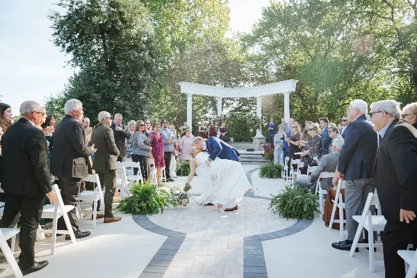 A bride and groom sharing a kiss during an outdoor wedding ceremony in a wooded area with a white arched structure and guests seated in white chairs.