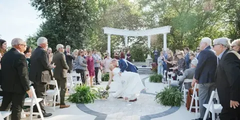 A bride and groom sharing a kiss during an outdoor wedding ceremony in a wooded area with a white arched structure and guests seated in white chairs.