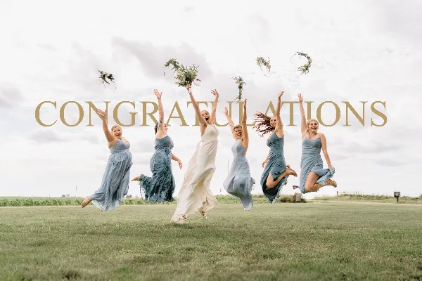 A bride and five bridesmaids in matching dresses jumping in a grassy field while tossing bouquets into the air.