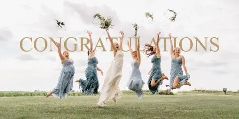 A bride and five bridesmaids in matching dresses jumping in a grassy field while tossing bouquets into the air.
