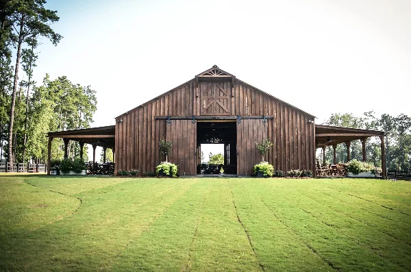 A large wooden barn with open doors and covered outdoor seating areas on a green lawn.