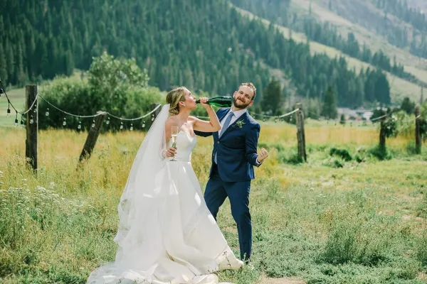 A bride and groom sharing a champagne toast in a grassy field with a mountainous backdrop.