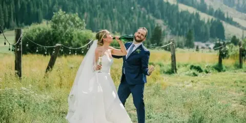 A bride and groom sharing a champagne toast in a grassy field with a mountainous backdrop.