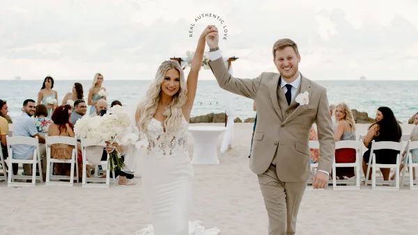 A bride and groom walking down a beach wedding aisle with seated guests and the ocean in the background.
