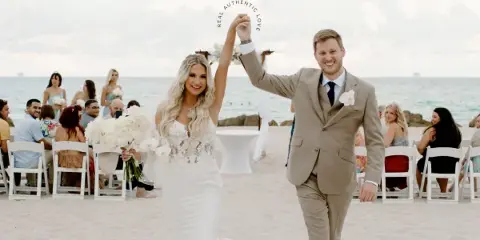 A bride and groom walking down a beach wedding aisle with seated guests and the ocean in the background.