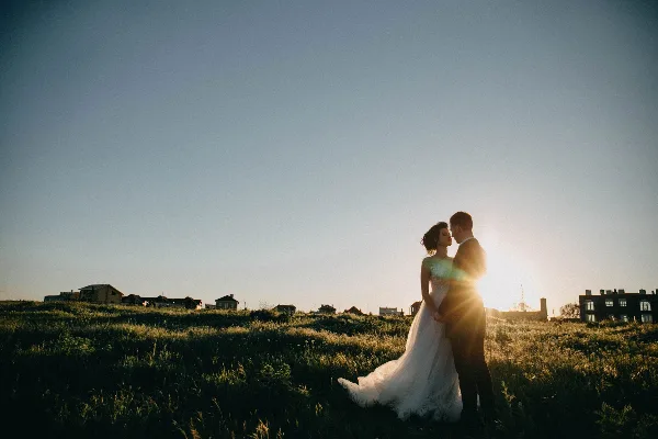 A bride and groom embracing in a grassy field at sunset.