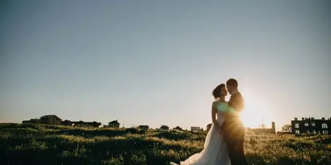 A bride and groom embracing in a grassy field at sunset.