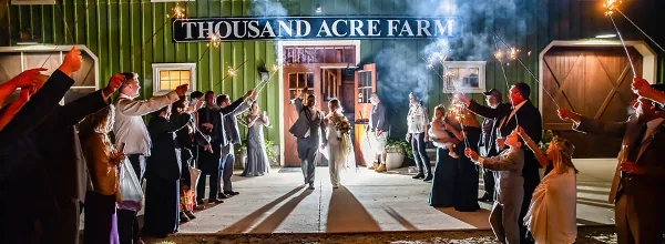 A wedding couple walks out of a barn venue at night while guests line both sides holding lit sparklers.