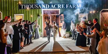 A wedding couple walks out of a barn venue at night while guests line both sides holding lit sparklers.