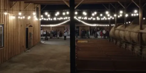 Interior view of a wooden barn venue with string lights and rows of benches set up for a wedding event.