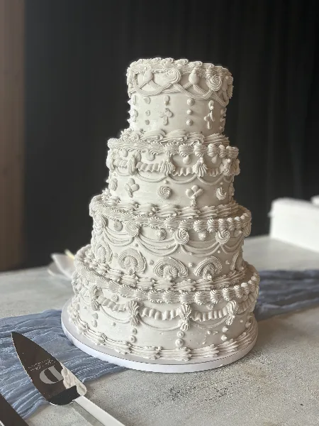 A four-tiered white wedding cake with intricate piped floral and scroll designs, sitting on a table with a cake server.