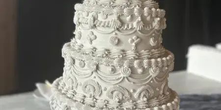 A four-tiered white wedding cake with intricate piped floral and scroll designs, sitting on a table with a cake server.