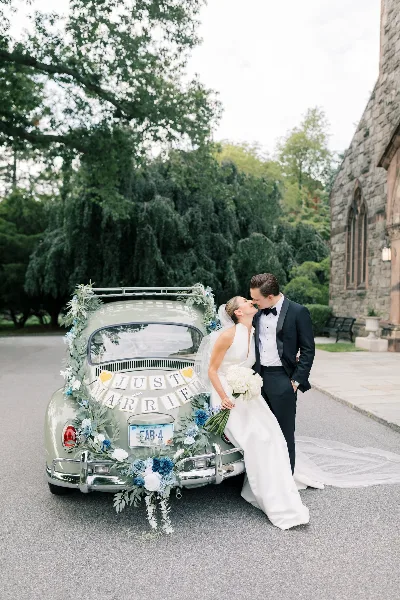 A bride and groom kissing next to a vintage car decorated with flowers and a 'Just Married' sign.