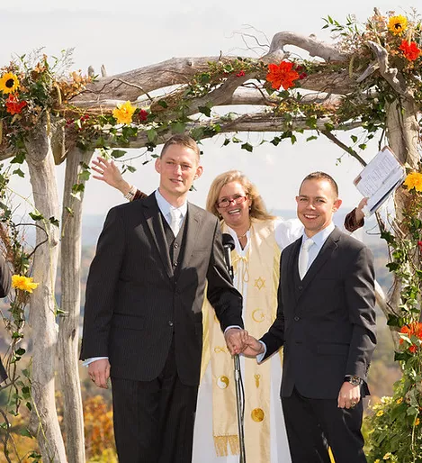 A female officiant in a white robe stands between two men in black suits at a wedding ceremony, holding their hands and speaking into a microphone.