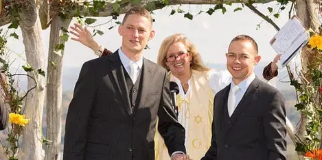 A female officiant in a white robe stands between two men in black suits at a wedding ceremony, holding their hands and speaking into a microphone.
