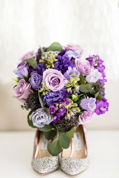 A purple and lavender bridal bouquet featuring roses, stock, and lisianthus resting on a pair of glittering high heels.