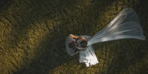 Aerial view of a bride and groom embracing in a grassy field, with the bride's veil flowing in the wind.
