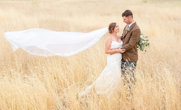 A bride and groom standing in a field of tall grass, with the bride's veil blowing in the wind.