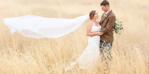 A bride and groom standing in a field of tall grass, with the bride's veil blowing in the wind.