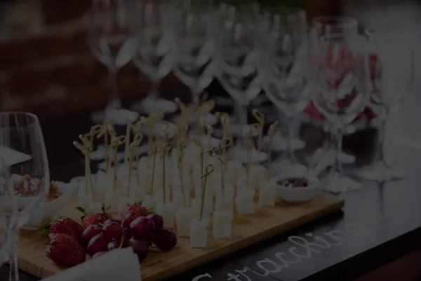 A catering table featuring a wooden board with cheese cubes on skewers, strawberries, and grapes, with wine glasses in the background.