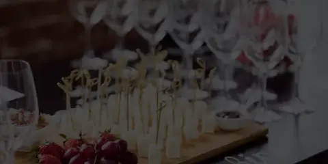 A catering table featuring a wooden board with cheese cubes on skewers, strawberries, and grapes, with wine glasses in the background.