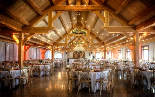 Interior view of a large timberframe banquet hall with exposed wooden beams, string lights, and round tables set for a wedding reception.