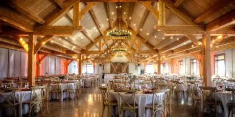 Interior view of a large timberframe banquet hall with exposed wooden beams, string lights, and round tables set for a wedding reception.