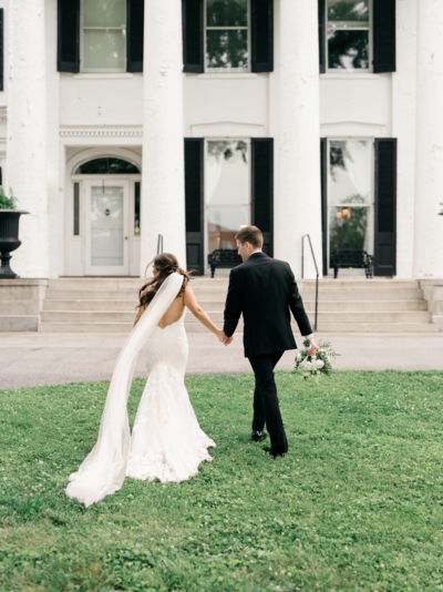 Bride in a white gown and groom in a black suit walking hand-in-hand across a lawn in front of a white building.
