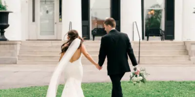 Bride in a white gown and groom in a black suit walking hand-in-hand across a lawn in front of a white building.