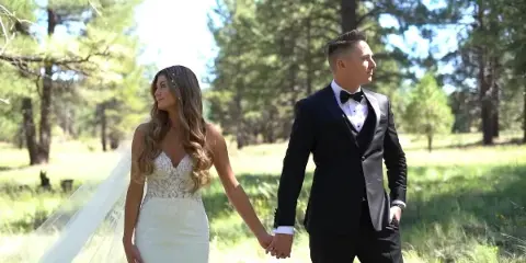 A bride and groom holding hands in a grassy outdoor area with trees in the background.