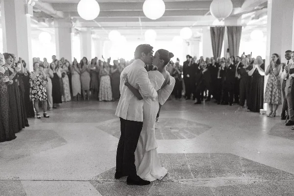 A black and white photograph of a bride and groom sharing a first dance on a reception floor while surrounded by guests.